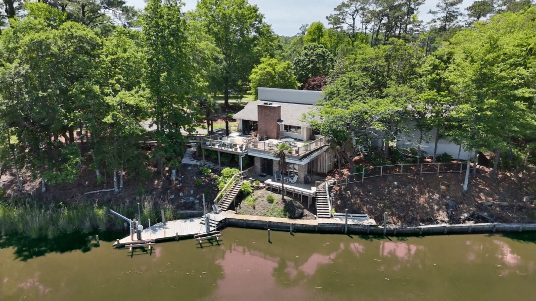 A lakeside house with multiple decks, surrounded by tall trees, and a wooden dock along a calm river.